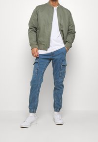 Chaqueta bomber verde, camiseta blanca, pantalones cargo azules con bolsillos y zapatillas blancas. Conjunto casual con un corte relajado y puños elásticos.