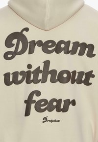 Beige Kapuzenpullover mit großen schwarzen Schriftzug "Traum ohne Angst", der prominent auf dem Rücken platziert ist, sowie einem Logo in der Nähe des unteren Bereichs.