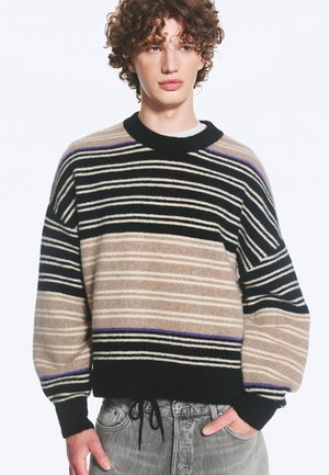 Jeune personne aux cheveux bouclés portant un pull rayé noir, beige et violet et un jean gris clair, une main dans la poche.