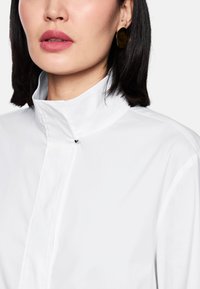 White high-collar shirt with a textured fabric, a hidden button closure, and a small heart-shaped detail on the front, paired with large round earrings.