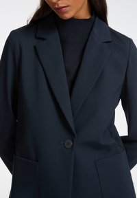 Granatowa marynarka uszyta z gładkiego materiału, z pojedynczym guzikiem, klapami w stylu notched oraz dwiema kieszeniami z przodu. Miękki, zdefiniowany krój.