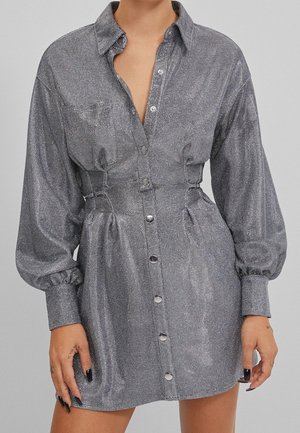 Langärmliges, silberfarbenes Mini-Kleid mit Metallknöpfen, geraffter Taille und Bündchen, getragen von einer Person mit schwarzem Nagellack und kleinen Handtattoos.