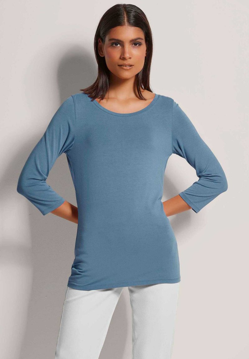 Langärmliges Oberteil aus hellblauem Stoff mit runder Ausschnitt und Dreiviertelärmeln, kombiniert mit weißen Hosen. Glatte Textur.