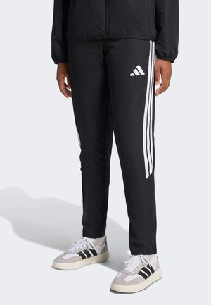 Person trägt schwarze Adidas-Trainingshose mit weißen Streifen und Logo, kombiniert mit weißen Sneakers mit schwarzen Streifen.