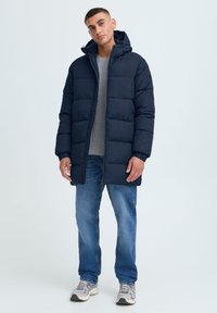 Mørkeblå puffjakke med hette, med kviltet design, frontglidelås og elastiske mansjetter, båret over en grå genser og blå jeans.