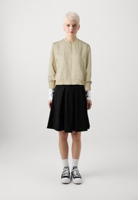 Béžová bomber bunda se zlatým zipem, spárovaná s černou plisovanou sukní, bílými ponožkami a černými teniskami. Hladká tkanina s minimálním designem.