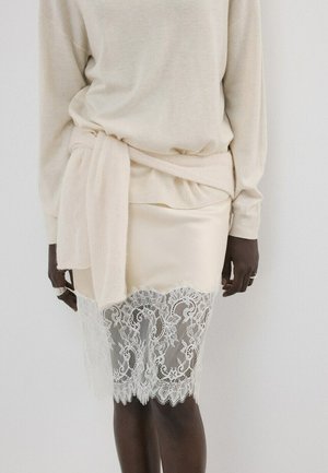 Persona que lleva un suéter crema con la cintura atada y una falda crema con un dobladillo de encaje floral sobre un fondo claro y liso.