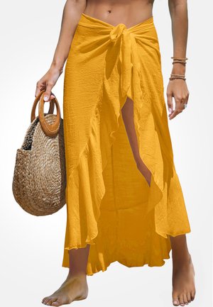 Gonna gialla avvolgente con fiocco frontale, orlo arricciato e spacco alto. Abbinata a una borsa in paglia intrecciata con manici in legno.