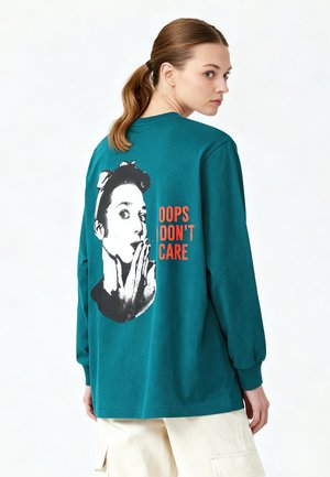 Junge Frau mit Pferdeschwanz, die ein türkisfarbenes Langarmshirt mit schwarz-weißem Aufdruck eines überraschten Gesichts und dem Text „OOPS I DON'T CARE“ trägt.