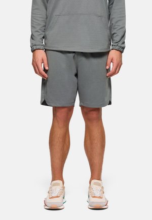 Hombre vistiendo una sudadera gris de manga larga con capucha y pantalones cortos a juego, con zapatillas blancas y beige, de pie contra un fondo blanco.