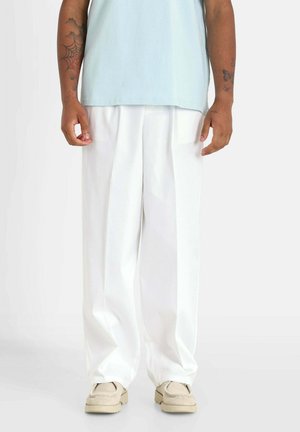 Osoba nosząca luźne białe spodnie, beżowe buty i jasnoniebieską koszulę, pokazująca tatuaże na obu przedramionach na jednolitym tle.