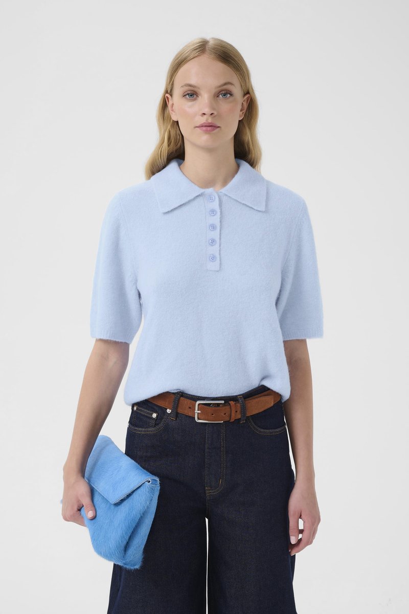 Frau trägt einen hellblauen Kurzarmpullover und dunkle Jeans mit braunem Gürtel, hält eine leuchtend blaue flauschige Clutch.