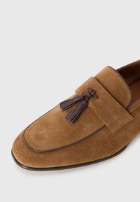 Hnedé semišové loafers s hladkou textúrou, doplnené koženou ozdobou v tvare strapca a kontrastným prešívaním pozdĺž okrajov.
