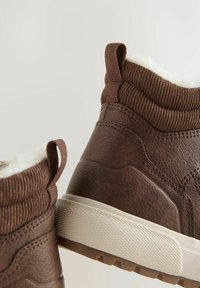 Braune Knöchelstiefel mit einer strukturierten Oberfläche, flauschiger Innenauskleidung und Ziehschlaufen, vor einem hellen Hintergrund präsentiert.