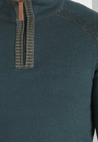 Maglione di lana color teal con collo alto e mezza cerniera. Cuciture strutturate vicino alla cerniera e tessuto del corpo liscio con una leggera lucentezza.