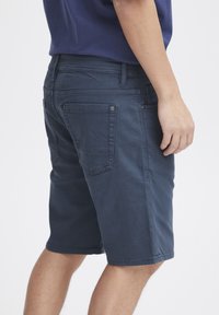 Temno modri bombažni šorts z oprijetim krojem, ki vključujejo hrbtne žepke in subtilne šive. Površina je gladka z rahlim sijajem.