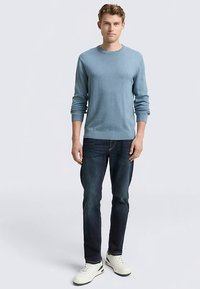 Hellblauer Pullover mit Rundhalsausschnitt, weicher Stoff, lockere Passform, kombiniert mit dunkelblauen Jeans und weißen Turnschuhen. Steht vor einfarbigem Hintergrund.