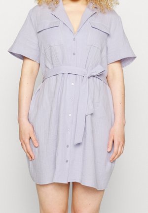 Vrouw draagt een lichtpaars, kort mouw button-up jurk met borstzakken en een gestrikte ceintuur in de taille, staand tegen een effen achtergrond.