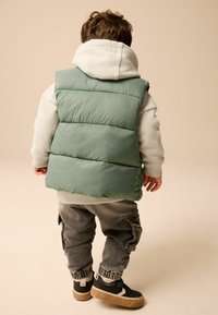 Zielona puchowa kamizelka, szara bluza z kapturem, szare spodnie cargo i czarne sneakersy. Kamizelka ma pikowany teksturę. Prosty, casualowy design.