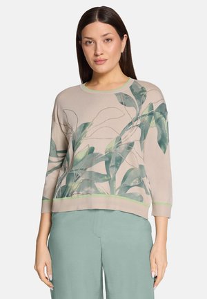 Vrouw met stijl donker haar draagt een beige trui met een groene bladprint en lichtgroene broek met wijde pijpen, staand tegen een witte achtergrond.