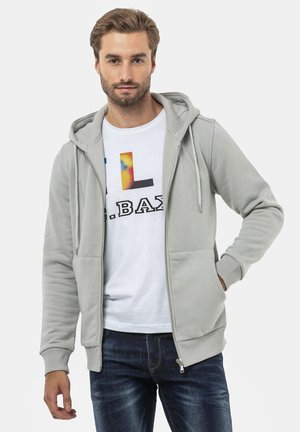 Haut en sweatshirt zippé gris clair avec capuche à cordon de serrage, présentant deux poches avant et des poignets côtelés, porté sur un t-shirt graphique blanc et un jean.