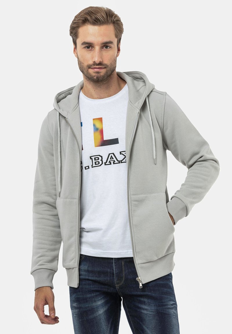 Haut en sweatshirt zippé gris clair avec capuche à cordon de serrage, présentant deux poches avant et des poignets côtelés, porté sur un t-shirt graphique blanc et un jean.
