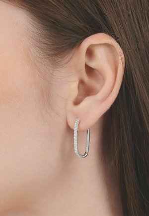 Primer plano de la oreja de una mujer con cabello liso y castaño, que lleva un pendiente de aro rectangular plata adornado con pequeñas piedras claras en un lado.