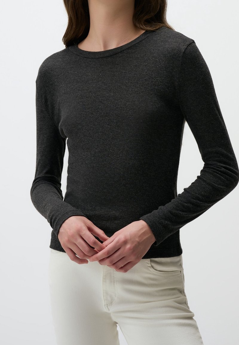 Femme portant un haut moulant à manches longues gris foncé et un pantalon blanc, debout les mains jointes devant un fond uni.