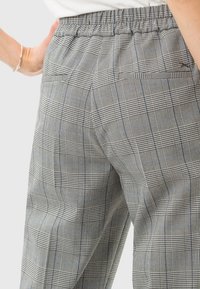 Pantalons à carreaux gris en tissu texturé, avec une taille élastique et des poches arrière. Le motif inclut des détails à carreaux noirs et bleus.