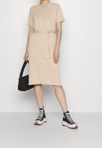 Beige mekko polven pituudelle, jossa on lyhyet hihat, kangashihna ja sileä pinta, yhdistettynä mustiin korkeatoppaisiin lenkkareihin ja mustaan laukkuun.