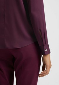 Langärmliges Hemd in tiefem Lila mit glatter Textur, featuring gespaltene Bündchen, verziert mit silbernen Knöpfen, kombiniert mit passenden Hosen.