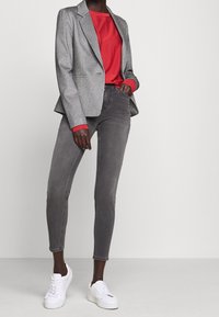 Šedé texturované sako přes červenou košili, doplněné šedými džíny a bílými teniskami. Důraz na přizpůsobený střih a kontrastní barvy.