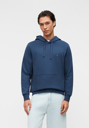 Giovane uomo in piedi che indossa una felpa con cappuccio blu con tasca frontale e jeans azzurri su uno sfondo neutro.