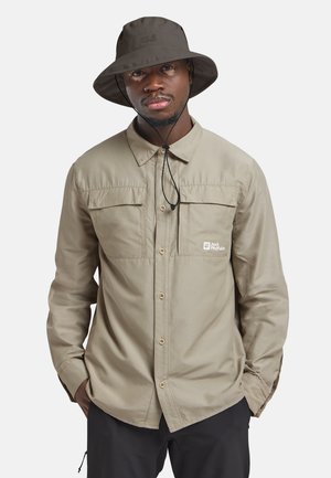 Homme portant une chemise kaki boutonnée et un chapeau d'extérieur sombre avec sangle sous le menton, les mains dans les poches, regardant légèrement devant avec une expression neutre.