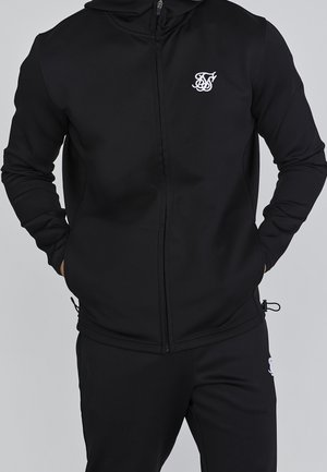 Mann trägt schwarzen Kapuzenpullover mit Reißverschluss und weißem Logo auf der Brust sowie schwarze passende Hose mit weißem Logo am Oberschenkel, Hände in den Taschen.