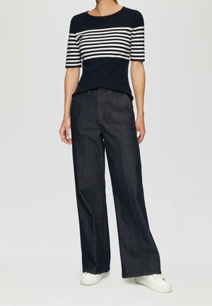 Person trägt ein marineblau-weiß gestreiftes kurzärmeliges Oberteil, weite dunkelblaue Denim-Hosen und weiße Turnschuhe, steht vor einem einfarbigen Hintergrund.