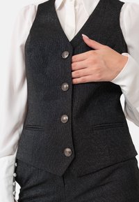 Zwarte gestructureerde kostuumvest met vijf zilveren knopen en zijzakken, gelaagd over een witte shirt met knoopaccenten op de manchetten.