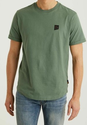 Mężczyzna noszący prostą oliwkowozieloną koszulkę z krótkim rękawem z małym kwadratowym logo na piersi oraz niebieskie dżinsy.