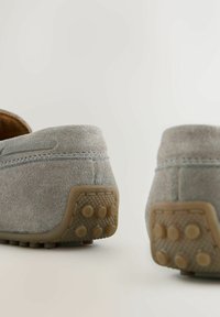 Pereche de mocasini din piele întoarsă gri deschis, cu tălpi de cauciuc cu textură de pietricele, văzuți din spate pe un fundal simplu, deschis la culoare.