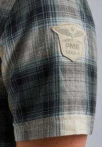 Korte mouwen plaid shirt in zwart, grijs en wit met een crème patch met vleugels en "PME 1649-A" op de bovenarm. Katoenen stof.