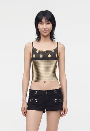 Mladi žena s ravnom crnom kosom nosi prugasti smeđe-bež bezrukavi crop top i crne kratke hlače sa srebrnim detaljima očnih jabučica.