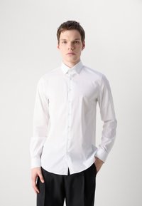Weißes Longsleeve-Shirt mit klassischem Kragen, Knopfverschluss und glatter Textur. Kombiniert mit schwarzen Hosen, minimale Markenkennzeichnung sichtbar.