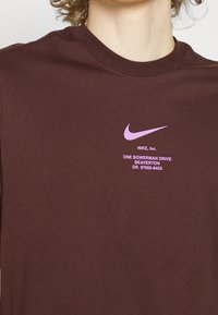 Bordowa koszulka wykonana z bawełny, z jasnofioletowym logo Nike oraz tekstem zawierającym dane adresowe, okrągły dekolt i krótkie rękawy.