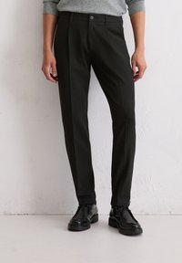 Pantalons noirs sur mesure en tissu léger avec une texture lisse, dotés d'une fermeture à bouton et d'une coupe slim. Portés avec des chaussures noires.
