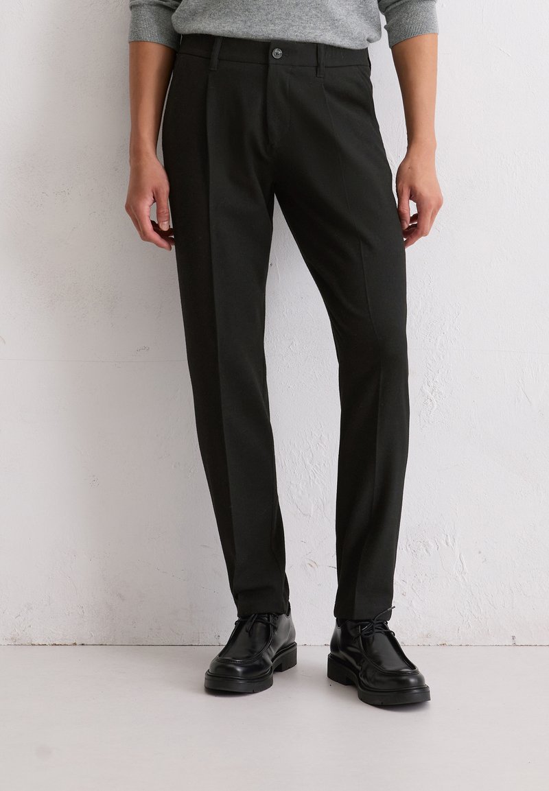 Pantalons noirs sur mesure en tissu léger avec une texture lisse, dotés d'une fermeture à bouton et d'une coupe slim. Portés avec des chaussures noires.