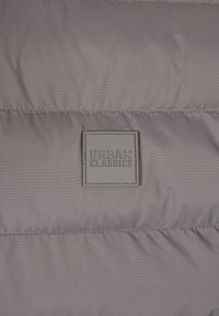 Grå pufferjakke materiale med quiltet tekstur, der har et præget firkantet mærke med "URBAN CLASSICS" logo. Synlige sømme.