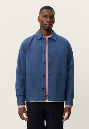 Blå bomuldsjakke med knaplukning foran, spids krave, to forlommer og en kontrasterende lyserød sweater under. Har lange ærmer.