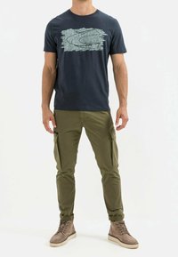 Marineblauw katoenen t-shirt met abstract teal graphic; olijfkleurige cargobroeken met zijzakken; lichtbruine hoge sneakers met beige zolen.