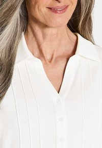 Blusa bianca realizzata in tessuto morbido con pieghe verticali, colletto e chiusura a bottoni. Presenta un collo a V e una texture liscia.