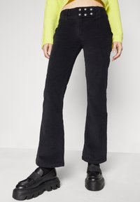 Pantalons évasés en velours côtelé noir avec une finition texturée et une taille haute, présentant trois boutons ronds sur la ceinture.